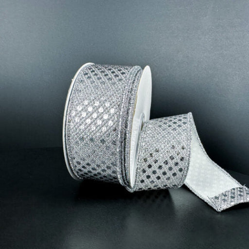 A spool of 1.5X10Y Silver Metallic Dragonscale Dots Ribbon (78521-09-51) rests on a dark surface, its sparkling, diamond-patterned length unrolled and curling forward.