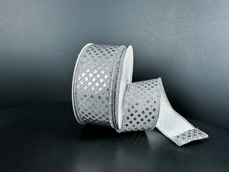 A spool of 1.5X10Y Silver Metallic Dragonscale Dots Ribbon (78521-09-51) rests on a dark surface, its sparkling, diamond-patterned length unrolled and curling forward.