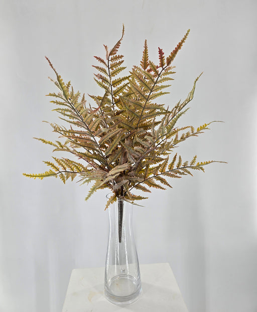 A clear glass vase holds multiple stems of the 21 Autumn Fern Bush 85360-UV-AUTUMN with yellow and brown leaves, displayed on a white surface against a plain white background.