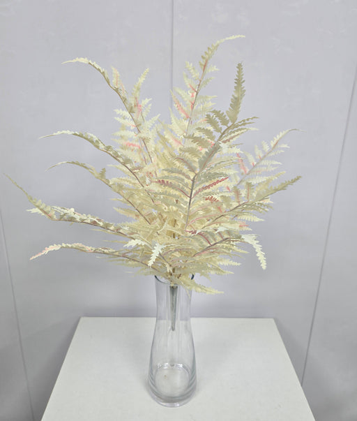 The 21 Cream Fern Bush (85360-UV-CR) with delicate, fern-like cream leaves is displayed in a clear glass vase on a light surface, set against a plain light gray wall.