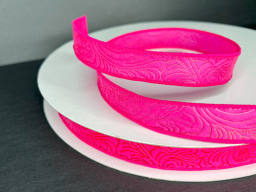 A 7/8X50Y (42466-05-28C) fuchsia flower-embossed ribbon, partially unrolled, sits on a white spool against a dark background. Its intricate swirling patterns enhance the elegant design.