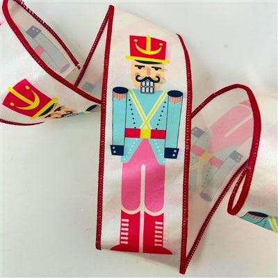 The 2.5" x 10 Pastel Nutcracker on Satin Ribbon (DCR5831) features a colorful soldier with a red hat, blue jacket, pink pants, and red boots, bordered with red trim on white satin.