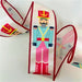 The 2.5" x 10 Pastel Nutcracker on Satin Ribbon (DCR5831) features a colorful soldier with a red hat, blue jacket, pink pants, and red boots, bordered with red trim on white satin.
