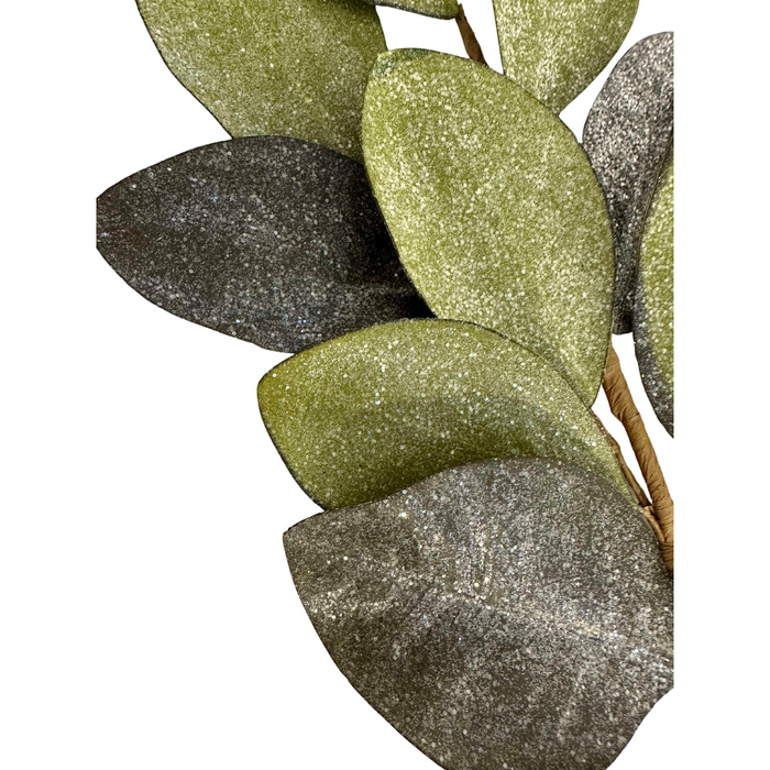 A close-up of the 30" Vintage Glitter Magnolia Leaves Spray ISB73133 showcases textured, glittery green and dark gray leaves arranged against a white background, adding elegance to any decor.