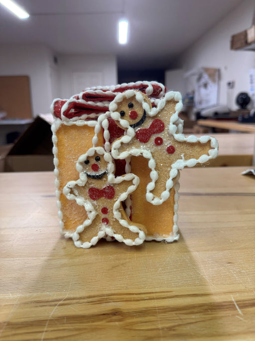 The 5" Gingerbread Gift Box Ornament (MTX75883) is shaped like a present and topped with two smiling gingerbread people decorated with white and red icing, displayed on a wooden table in a brightly lit room.