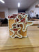 The 5" Gingerbread Gift Box Ornament (MTX75883) is shaped like a present and topped with two smiling gingerbread people decorated with white and red icing, displayed on a wooden table in a brightly lit room.