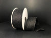 A spool of 1.5X10Y Black All Flat Glitter Ribbon (X820609-21) is shown against a black background, with part of the sparkling ribbon unrolled and draped in front.