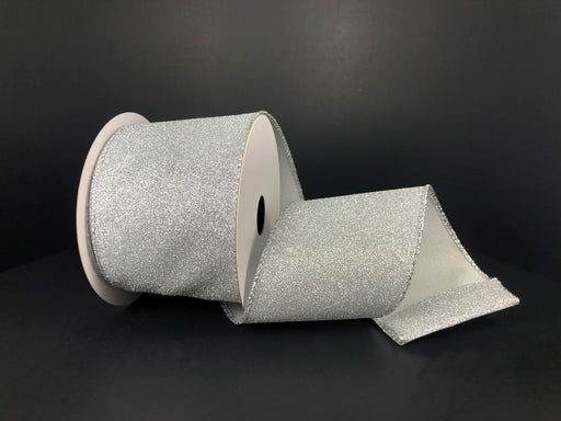 A 2.5X10Y Silver All Flat Glitter Ribbon X820640-16 with a white reel rests on a black surface, partially unrolled to display its sparkling flat glitter texture and finished edges.