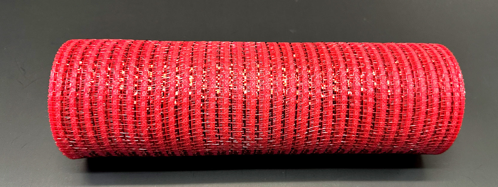 A roll of Red Breeze Metallic Mesh XB240810-12 measuring 10 inches by 10 yards lies horizontally on a smooth, dark surface. The woven texture of the mesh and its vibrant metallic red color create a striking contrast with the background.