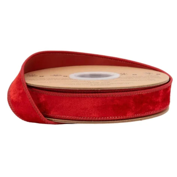 A spool of Heritage Red Velvet with Red Back Wired Ribbon (1 Inch x 10 Yards | 7005W-250-25E) is partially unwound, revealing its soft, shiny red velvet texture. The ribbon, featuring a cardboard center with a circular opening, is displayed against a plain white background.