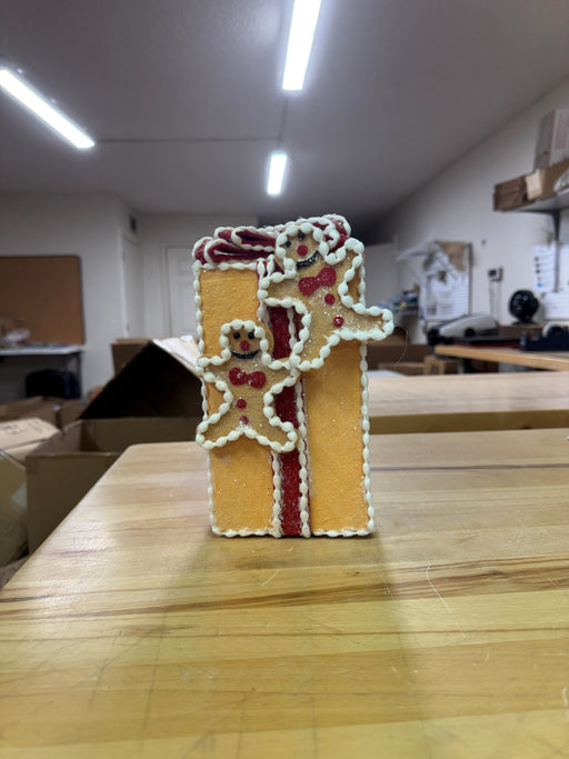 An 8" Gingerbread Gift Box Ornament (MTX75884), shaped like a yellow rectangle with a red ribbon and two smiling gingerbread people, sits on a wooden table in a room with shelves and boxes in the background.