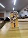 An 8" Gingerbread Gift Box Ornament (MTX75884), shaped like a yellow rectangle with a red ribbon and two smiling gingerbread people, sits on a wooden table in a room with shelves and boxes in the background.
