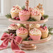 Flat metal Christmas sign featuring a distressed pink two-tiered tray filled with cupcakes decorated in pink and white frosting, Christmas sprinkles, peppermint candies, and a gingerbread star. Evergreen sprigs, frosted red berries, and a large pink velvet bow with gold trim accent the festive display. TTE-860.