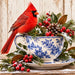 Flat metal winter sign featuring a bright red cardinal perched on a blue and white chinoiserie floral teacup filled with evergreen branches, red berries, magnolia leaves, and a pinecone dusted with snow. A matching saucer rests on a rustic wooden surface against a distressed white background. TTE-866.