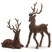 The Set of 2 Brown Flocked Christmas Deer (RAZ 4540866) features two charming brown deer figurines—one standing tall and the other lying down—each adorned with large antlers and crafted from a soft, velvety flocked material that evokes a classic woodland holiday look reminiscent of RAZ Imports’ signature style.