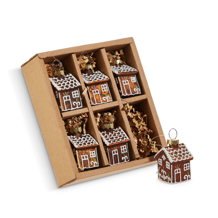 A cardboard box contains the Set of 6 Mini Gingerbread House Glass Ornaments (RAZ 4520866), each ornament beautifully crafted in the shape of a gingerbread house and adorned with delicate white icing designs. One charming glass gingerbread ornament is showcased outside the box, while brown paper filler keeps each ornament safely separated inside for protection.