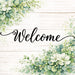 Welcome metal wreath sign with printed whitewashed plank background, green foliage and white flowers.