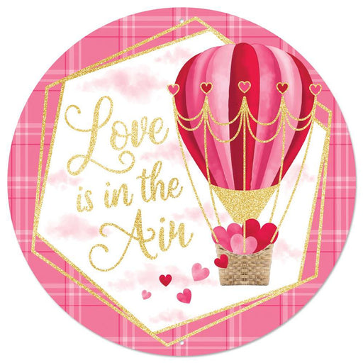 The 12Dia Metal/Foil Love Is In The Air Pink/Red/Hot Pink MD1073 sign features a round illustration of a red and pink heart-adorned hot air balloon beside the cursive Love Is In The Air in glittery gold. Small hearts float below on a soft pink plaid background.