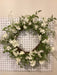 The 22 Cherry Blossom Wreath 62811, embellished with white flowers and green leaves, gracefully hangs on a white pegboard, enhancing any space with its floral charm.