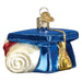 The Mortarboard 36254 Old World Christmas Ornament showcases a shiny blue graduation cap with a gold tassel and a sparkling rolled diploma tied with a red ribbon.